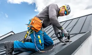 Dekarz w kasku i okularach montuje dachówkę wkrętarką.