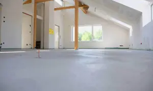 Nowa wylewka betonowa w stanie surowym, widoczne drewniane belki stropowe i okno.