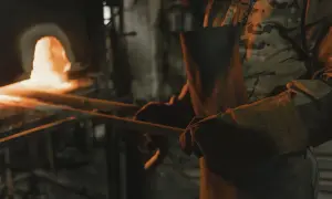 Osoba w fartuchu i rękawicach trzyma rozgrzany metal przy piecu.