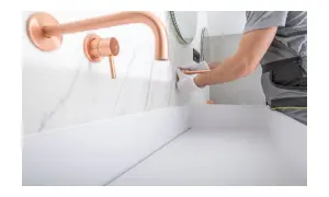 Instalator w białych rękawiczkach montuje miedzianą baterię podtynkową nad białym blatem.