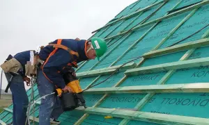 Dwaj dekarze pracują nad dachem, montując tymczasowe zabezpieczenie dachu. Jeden z nich używa pneumatycznego gwoździarki.