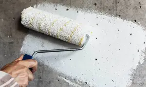Dłoń trzymająca wałek malarski z białą farbą na szarej, chropowatej ścianie.