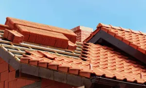 Układanie ceramicznych dachówek na dachu w budowie, widoczny komin i niebo.