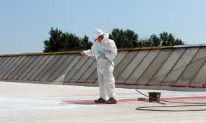 Pracownik w białym kombinezonie natryskuje białą papę na dach.