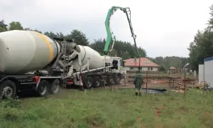 Betoniarka z wysięgnikiem pompującym beton na placu budowy, w tle dom i drzewa.