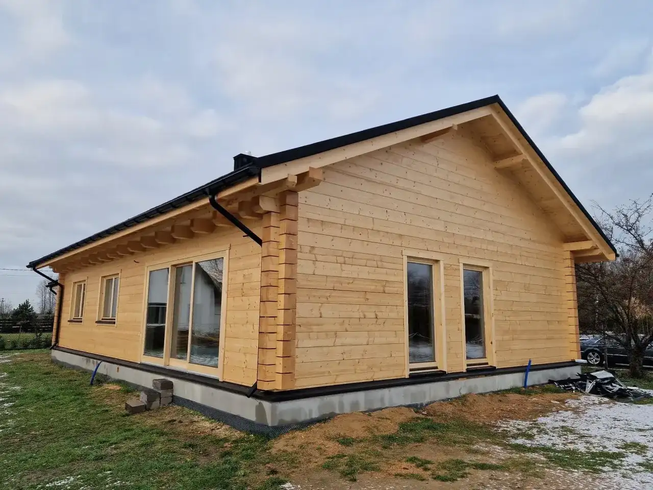 Nowy, parterowy dom z drewna klejonego z dużymi oknami i czarnym dachem, otoczony zielenią i resztkami śniegu.