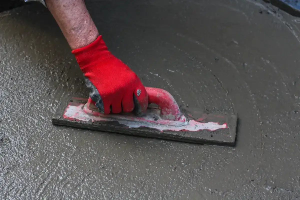 Ręka w czerwonej rękawicy wygładza cienką wylewkę na starym betonie za pomocą kielni.
