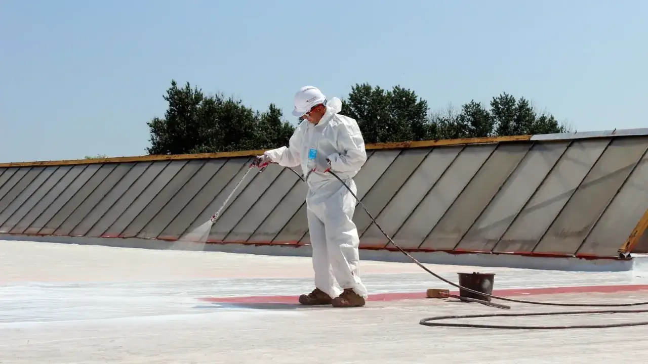 Pracownik w białym kombinezonie natryskuje białą papę na dach.