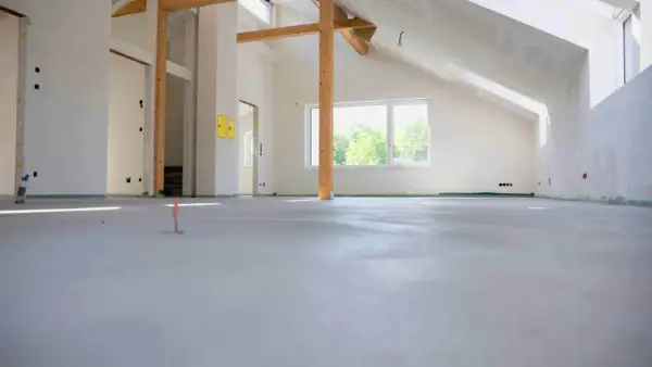Nowa wylewka betonowa w stanie surowym, widoczne drewniane belki stropowe i okno.