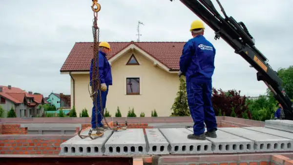 Dwóch budowlańców w żółtych kaskach montuje prefabrykowane płyty stropowe.