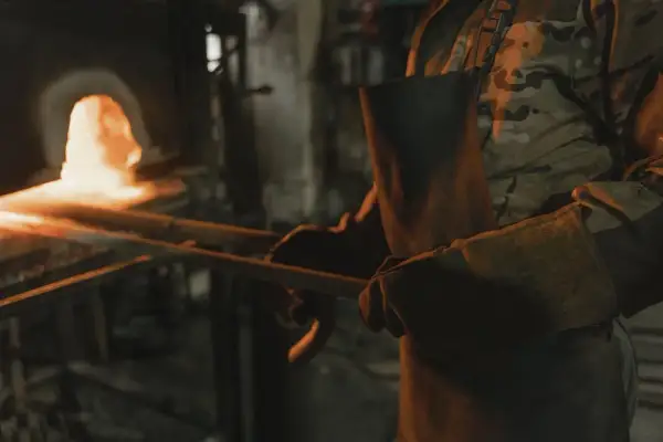 Osoba w fartuchu i rękawicach trzyma rozgrzany metal przy piecu.