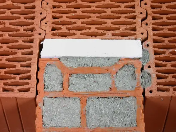 Zbliżenie na fragment ściany z pustaków ceramicznych, zaprawy i białego elementu izolacyjnego.