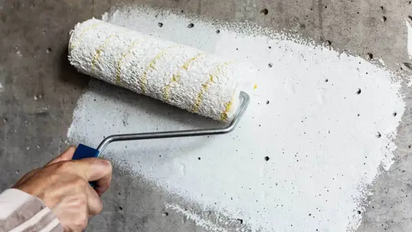 Dłoń trzymająca wałek malarski z białą farbą na szarej, chropowatej ścianie.