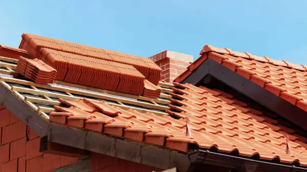Układanie ceramicznych dachówek na dachu w budowie, widoczny komin i niebo.
