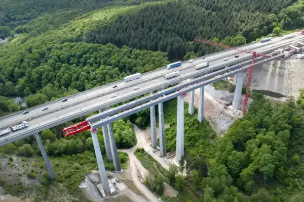 Widok z lotu ptaka na budowę betonowego mostu nad lasem, z ruchem samochodowym na ukończonej części.