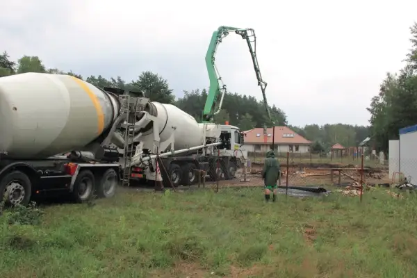 Betoniarka z wysięgnikiem pompującym beton na placu budowy, w tle dom i drzewa.