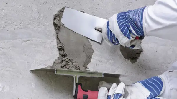 Dłonie w rękawicach nakładają zaprawę na betonową powierzchnię za pomocą kielni.