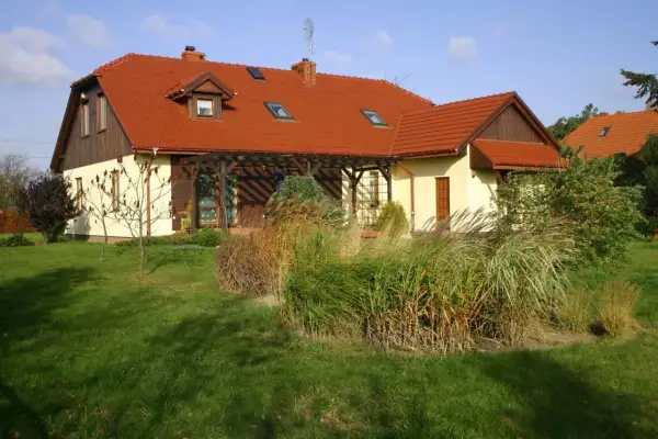 Dom z czerwonym dachem i naczółkami, otoczony zielonym trawnikiem i ozdobnymi trawami.